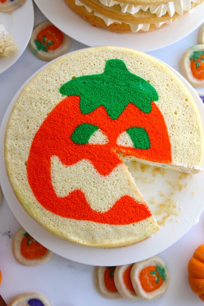 A round cake with a colorful Halloween-themed pumpkin design, featuring a green stem and orange face with a zigzag mouth. A slice is cut out, showing the vanilla layers inside. Small pumpkin-shaped cookies are scattered around.