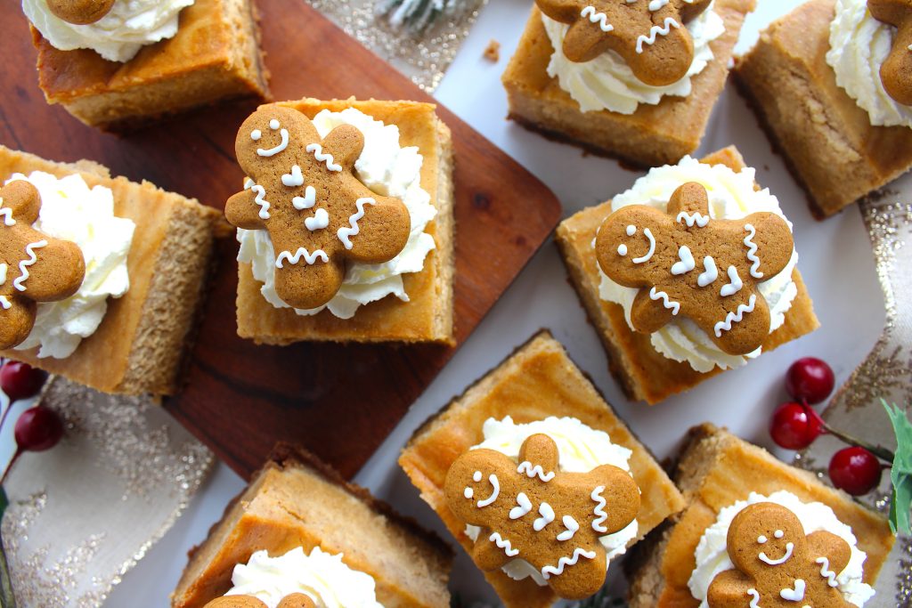 Square gingerbread cheesecake bars topped with whipped cream and small gingerbread cookies are arranged on a wooden board. Decorated with red berries and greenery, creating a festive holiday display.