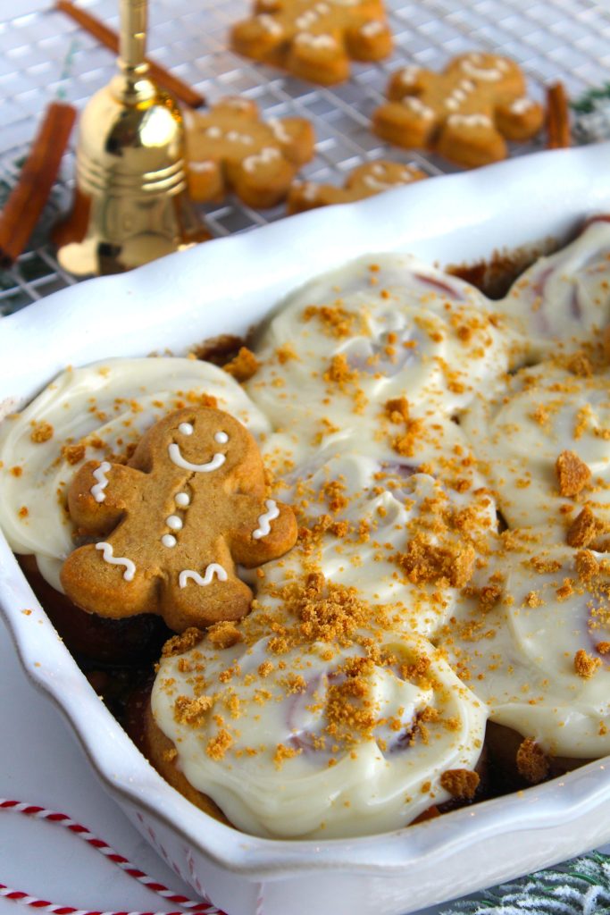 A festive dish of cinnamon rolls topped with white icing, garnished with crumbled gingerbread cookies, with a smiling gingerbread cookie placed on the side. A wire rack holds more gingerbread cookies in the background.