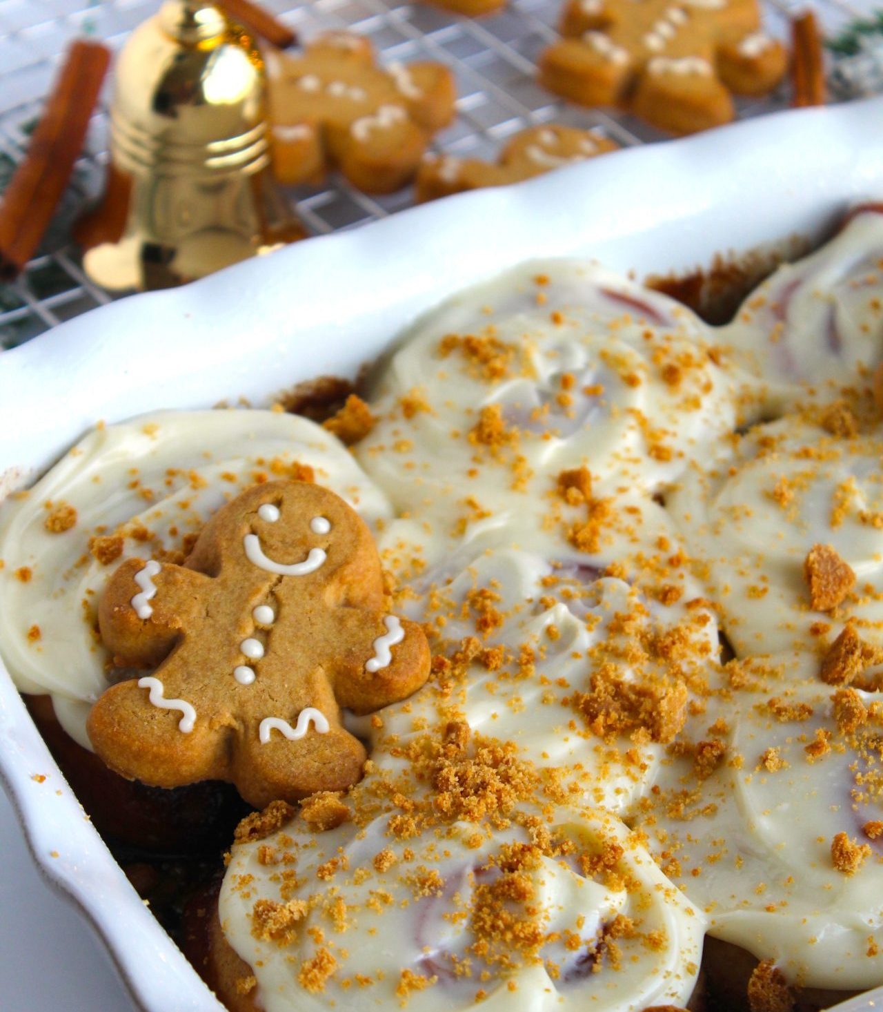 A festive dish of cinnamon rolls topped with white icing, garnished with crumbled gingerbread cookies, with a smiling gingerbread cookie placed on the side. A wire rack holds more gingerbread cookies in the background.