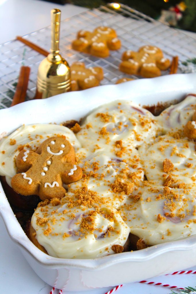A holiday-themed dessert in a white dish, topped with white icing and gingerbread cookie crumbs. A decorated gingerbread cookie is placed on top. In the background, a golden bell and more gingerbread cookies are on a cooling rack.