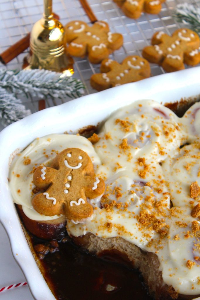 A festive dessert in a white dish topped with icing and a gingerbread cookie. Crumbled cookie pieces are sprinkled over the top. In the background, more gingerbread cookies are cooling on a rack, surrounded by holiday decorations.