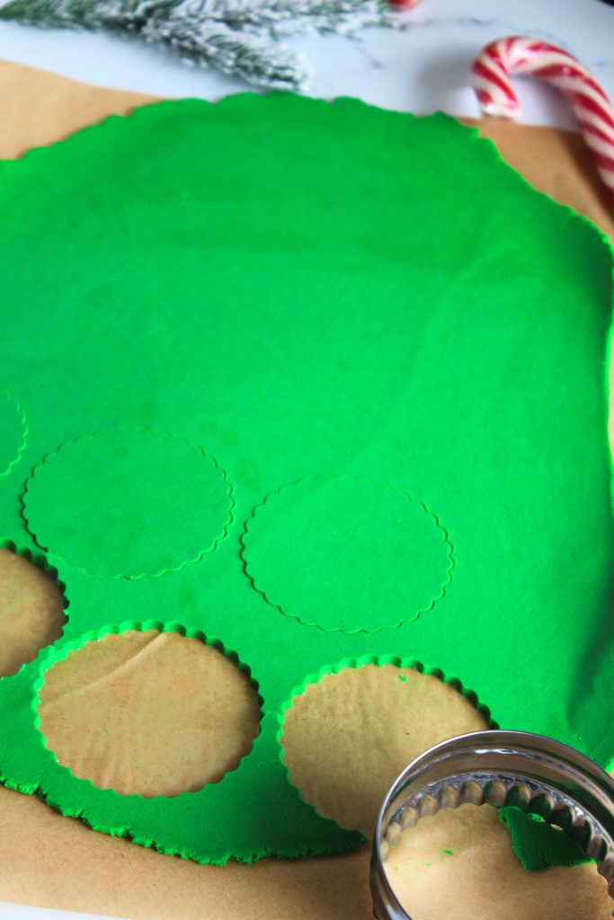 A rolled-out sheet of green cookie dough on parchment paper with circular shapes being cut out using a metal cookie cutter. A candy cane and pine branches are visible in the background.