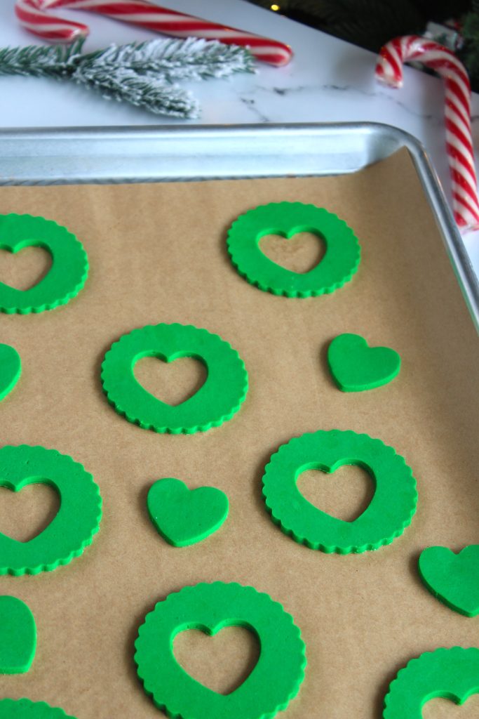 A baking tray lined with parchment paper, featuring several unbaked green cookies in circular shapes with a heart cutout in the center. There's a small heart-shaped cookie in the middle. Holiday decorations and a candy cane are in the background.