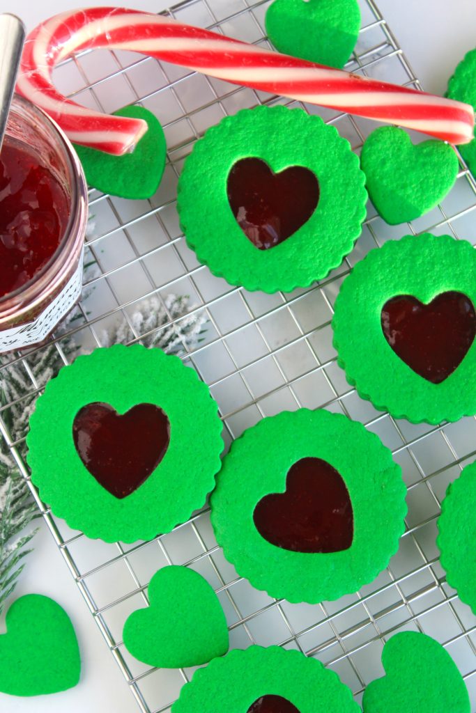 Green cookies with heart-shaped cutouts filled with red jam are displayed on a wire rack. A candy cane and a jar of red jam accompany the cookies, with pine sprigs in the background, creating a festive holiday atmosphere.