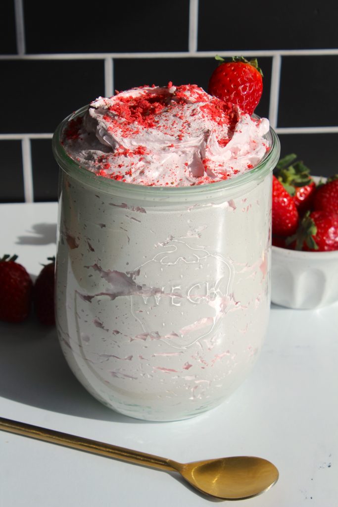 A glass jar filled with strawberry-flavored whipped cream topped with a strawberry, placed on a white surface. A gold spoon is beside the jar. A small bowl of fresh strawberries is in the background, against a black tiled wall.