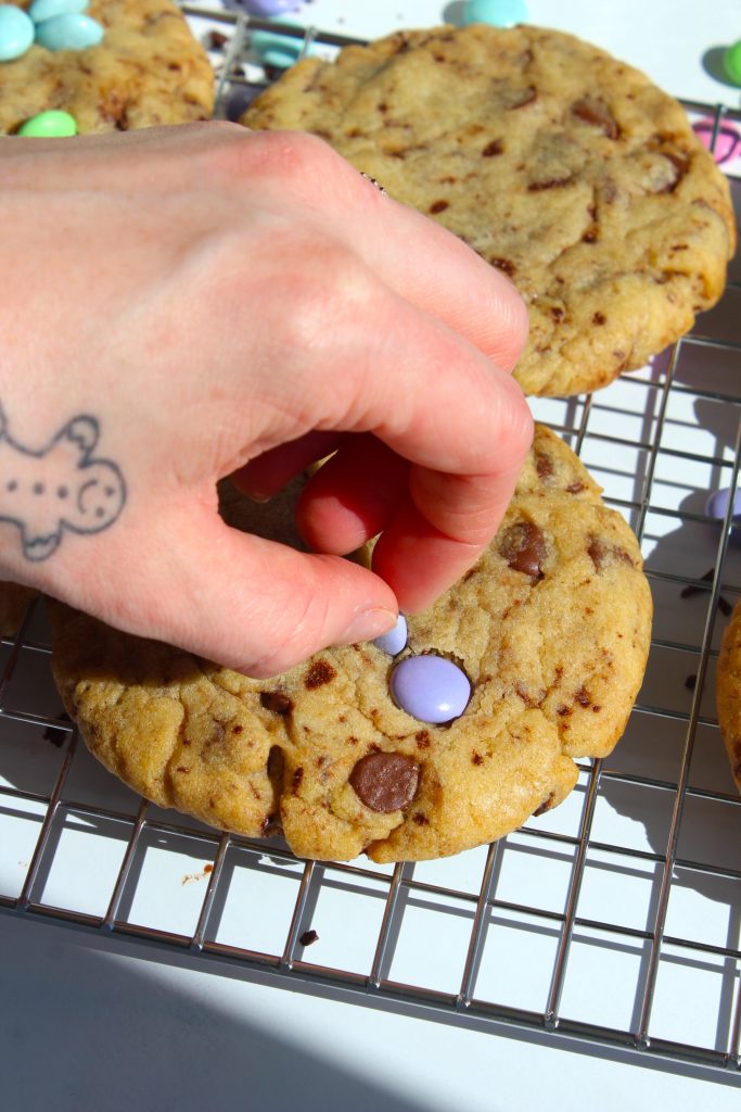 A hand with a gingerbread man tattoo places a pastel-colored candy on a chocolate chip cookie. The cookie rests on a wire cooling rack, surrounded by sunlight.