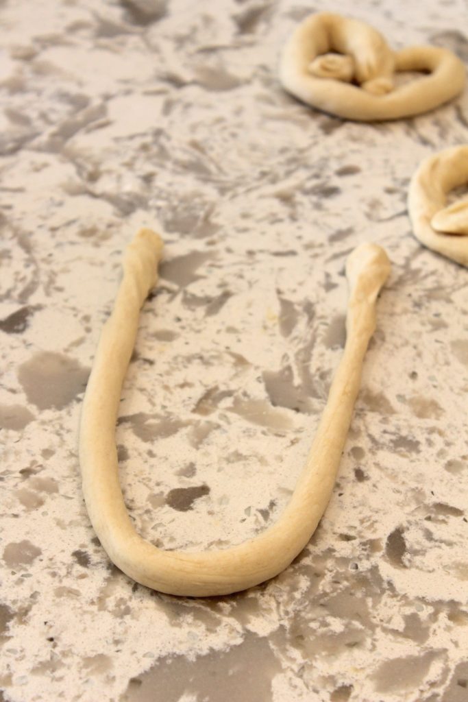 A piece of dough shaped like a horseshoe on a speckled countertop, with another similar-shaped dough in the background.