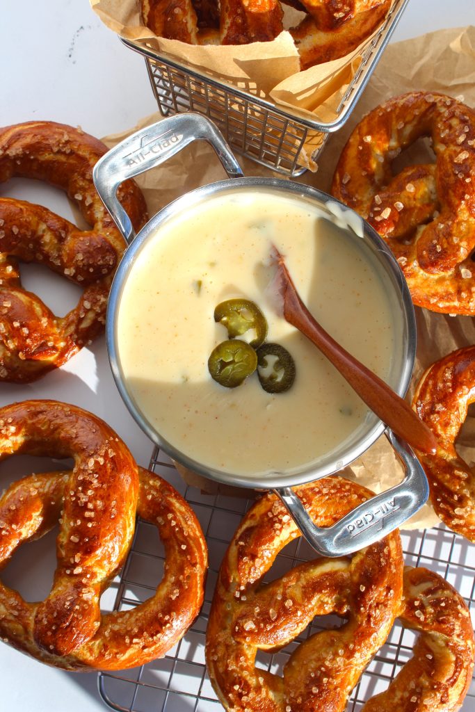 A table with freshly baked pretzels sprinkled with coarse salt, surrounding a pot of creamy cheese dip garnished with jalapeƱo slices. A wooden spoon rests inside the pot, ready for serving. A basket holds some pretzels in the background.