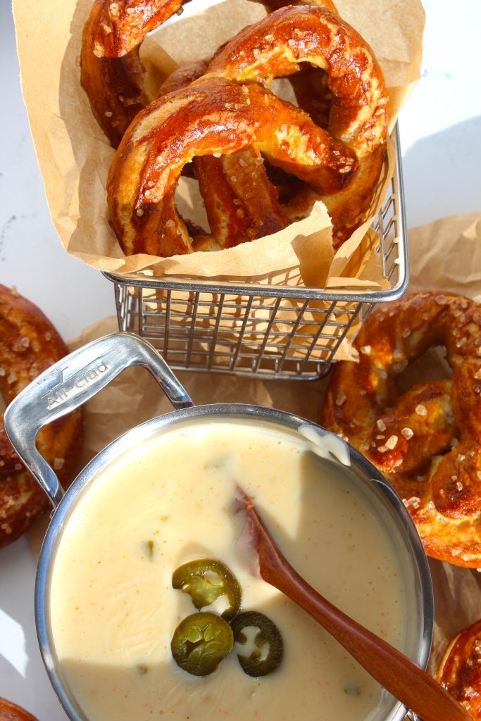 A basket of soft pretzels with a metal dish of cheese sauce in the foreground. The sauce is creamy with visible jalapeƱo slices, and a wooden spoon rests in it. The scene is well-lit, showcasing the pretzels' golden brown color.