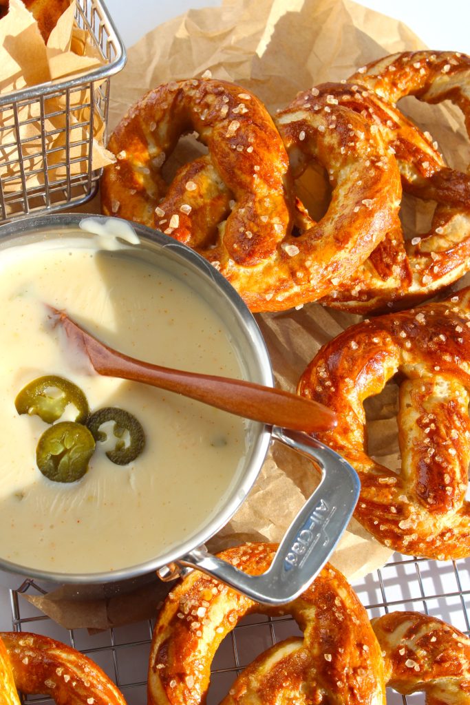 Golden-brown pretzels topped with coarse salt are arranged on parchment paper. A metal bowl of creamy cheese dip, garnished with jalapeƱo slices and a wooden spoon, is in the foreground. A small basket of parchment holds more pretzels.