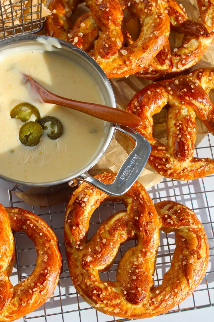 A collection of golden-brown pretzels sprinkled with coarse salt is laid on a cooling rack. A metal bowl filled with creamy cheese sauce, garnished with sliced jalapeños, is placed among the pretzels, with a wooden spoon resting in the bowl.