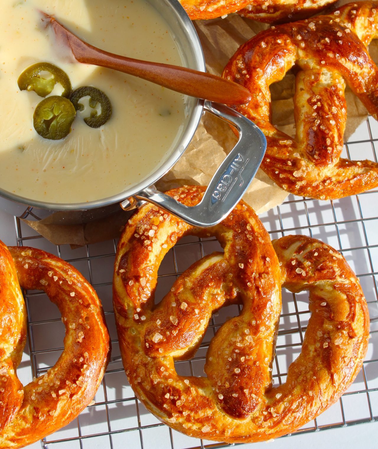 A collection of golden-brown pretzels sprinkled with coarse salt is laid on a cooling rack. A metal bowl filled with creamy cheese sauce, garnished with sliced jalapeƱos, is placed among the pretzels, with a wooden spoon resting in the bowl.