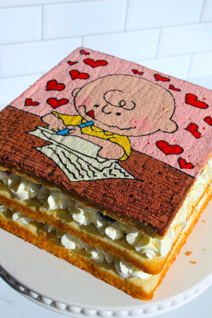 Square layered cake with white and chocolate cream between layers. The top layer features an illustration of a cartoon character wearing a yellow shirt, writing on paper, surrounded by red hearts against a pink background.