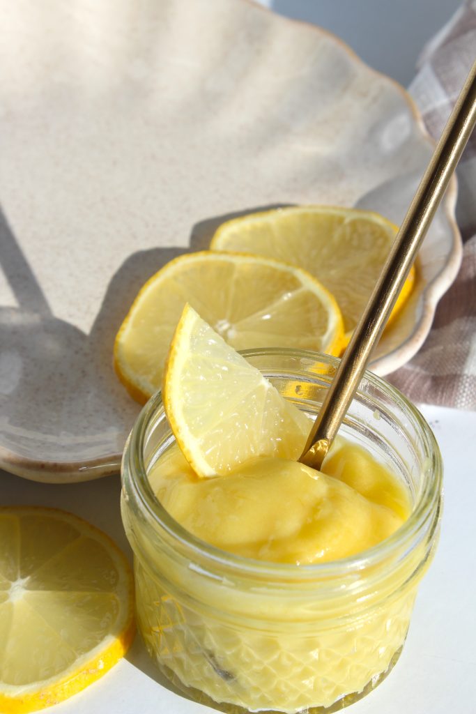 A small glass jar filled with creamy lemon curd, topped with a lemon slice and a gold spoon. Slices of lemon are arranged on a white, scalloped-edge plate in the background, casting soft shadows.