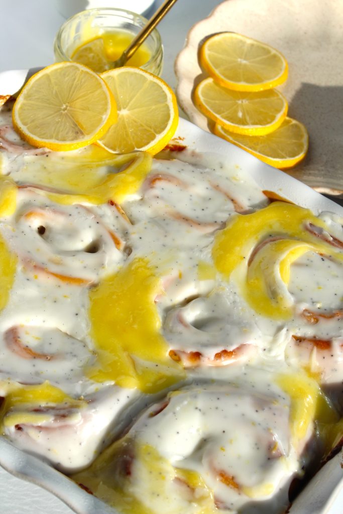 A close-up of lemon-glazed cinnamon rolls in a white baking dish. The rolls are topped with creamy icing and lemon curd. There is a small jar filled with lemon glaze and a spoon nearby, with lemon slices on a plate beside it.