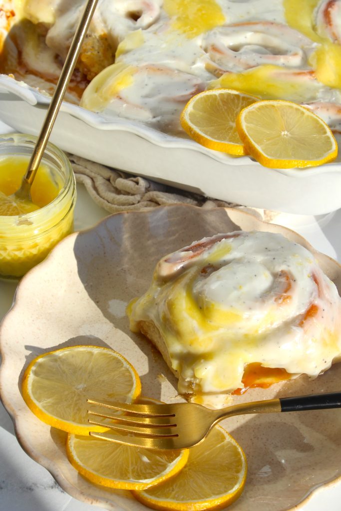 A plate with a frosted cinnamon roll and lemon slices. A fork rests nearby. A glass jar with lemon curd and a golden spoon sits next to a baking dish filled with frosted cinnamon rolls, topped with lemon slices. Sunlight casts soft shadows.