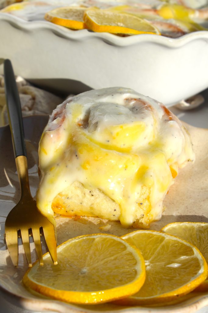 A close-up of a lemon cinnamon roll topped with creamy glaze on a plate. The roll is accompanied by sliced lemons, and a gold fork rests beside it. In the background, more pastries and lemon slices are visible.