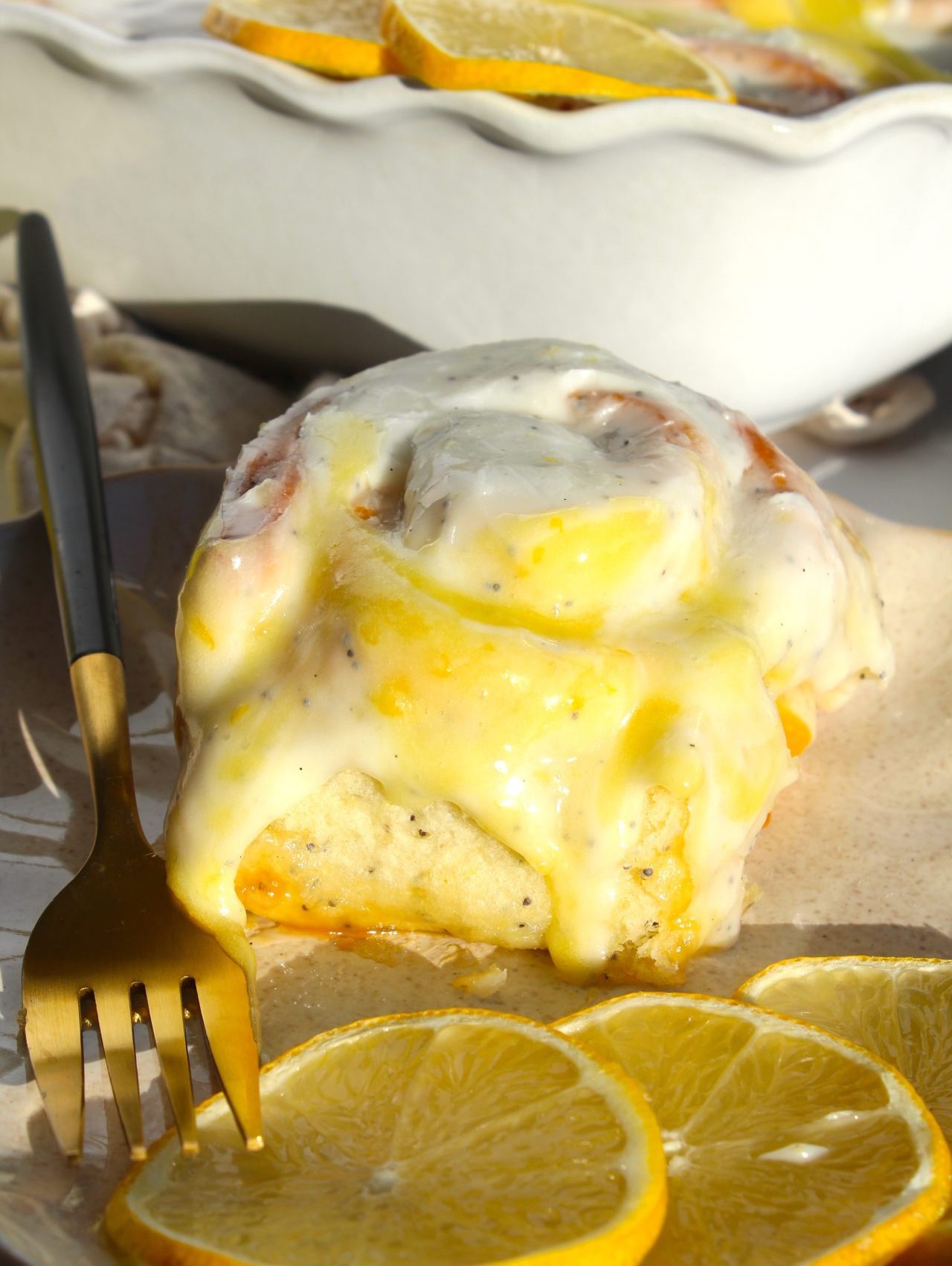 A close-up of a lemon cinnamon roll topped with creamy glaze on a plate. The roll is accompanied by sliced lemons, and a gold fork rests beside it. In the background, more pastries and lemon slices are visible.