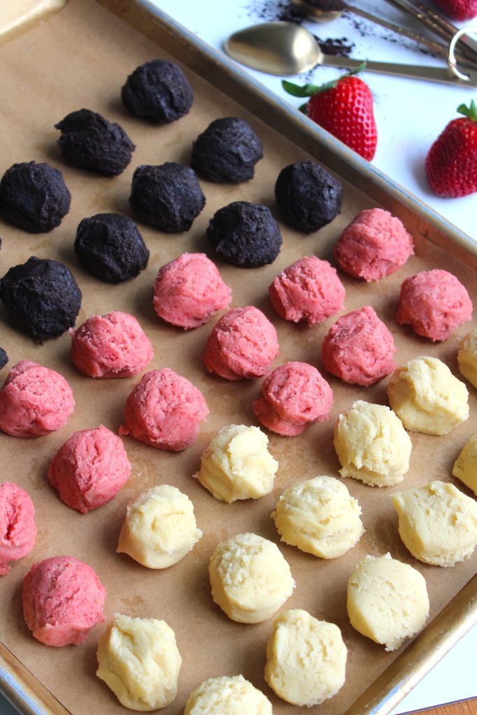 Rows of cookie dough scoops in three flavors: chocolate, strawberry, and vanilla, arranged on a parchment-lined baking sheet. Strawberries and measuring spoons are visible in the background.
