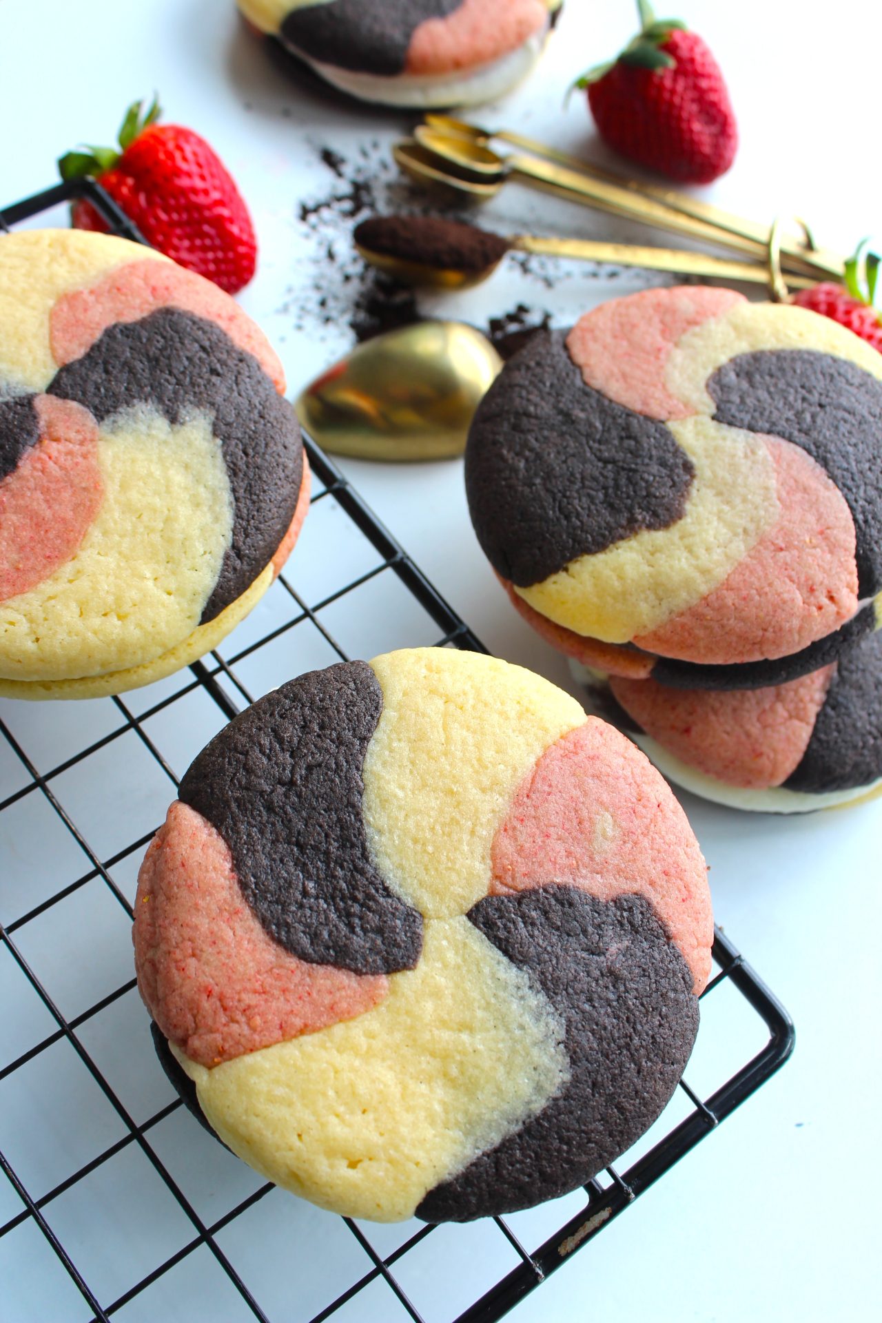 A close-up of cookies with a swirl pattern of brown, pink, and yellow portions, arranged on a cooling rack. Fresh strawberries and gold utensils are in the background, creating a colorful and appetizing display.