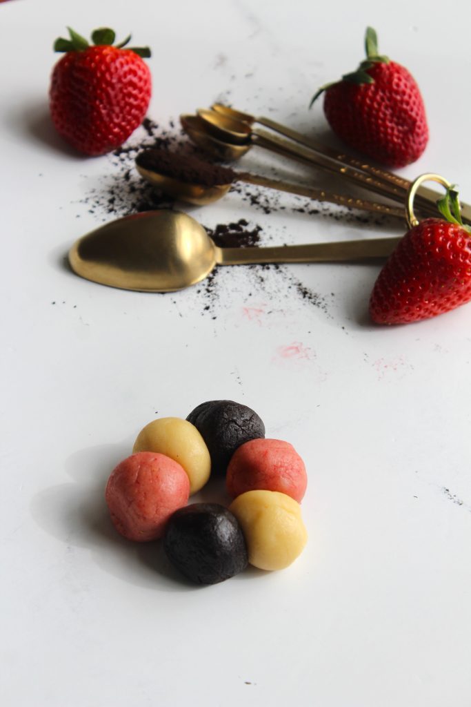 Assorted balls of dough in red, yellow, and black are arranged on a white surface. Behind them are three strawberries and four gold measuring spoons with cocoa powder scattered around.