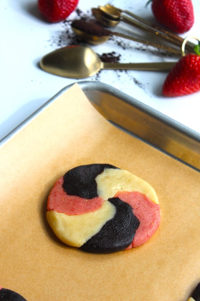 A swirled cookie with black, red, and yellow segments on a baking tray with parchment paper. Strawberries and measuring spoons filled with cocoa powder are in the background.