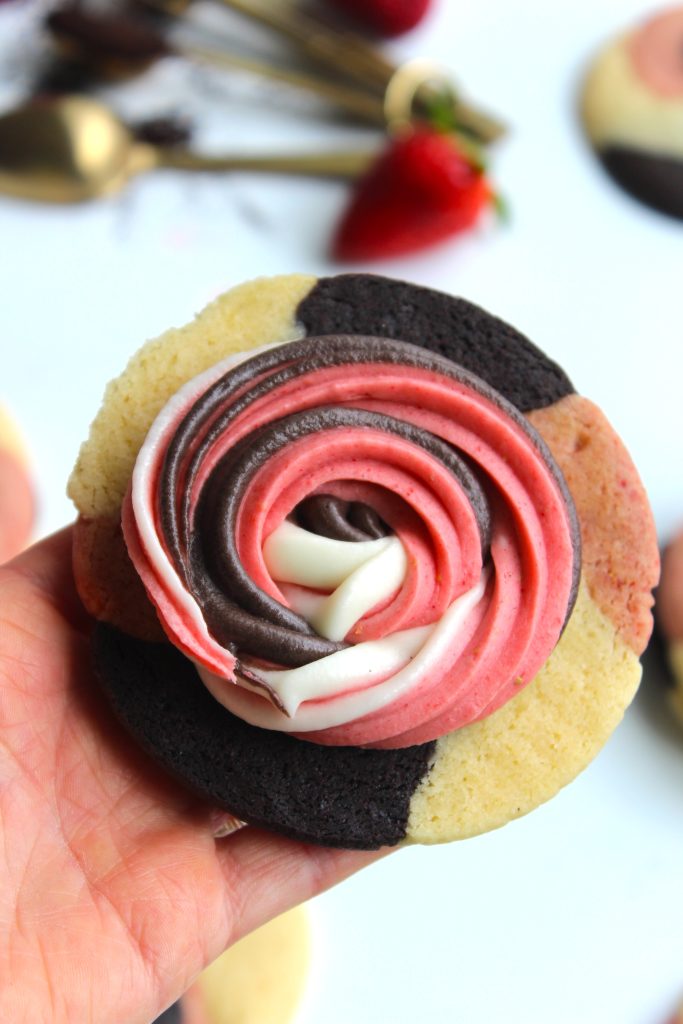 A hand holds a tricolor cookie with sections of vanilla, chocolate, and strawberry flavors. Swirled frosting on top matches the same three colors. Blurred strawberries and spoons are in the background.