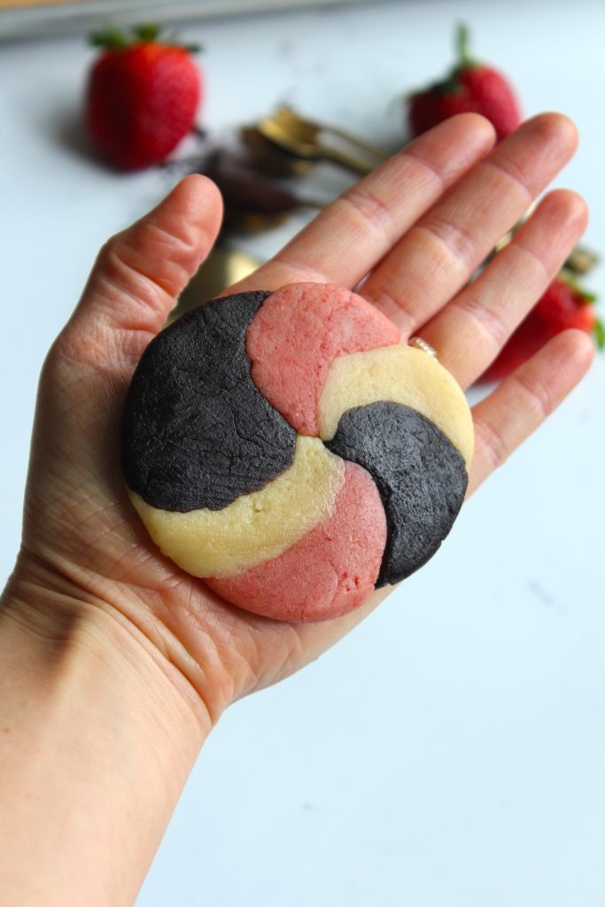 A hand holding a round, spiral-patterned cookie with three colored sections: black, pink, and cream. Blurred strawberries and spoons are in the background on a white surface.