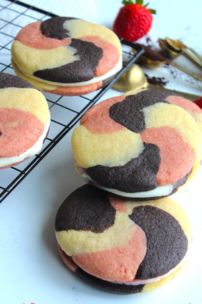 Four swirl cookies with chocolate, strawberry, and vanilla colors are stacked on a white surface, with one stack on a cooling rack. A strawberry, spoons, and cocoa powder are scattered nearby.