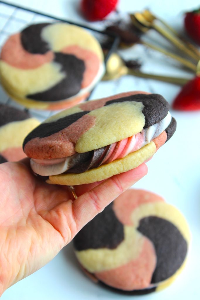 A hand holds a Neapolitan cookie sandwich filled with pink, white, and brown cream. The cookie features swirls of chocolate, vanilla, and strawberry dough. More cookie sandwiches and strawberries are blurred in the background.