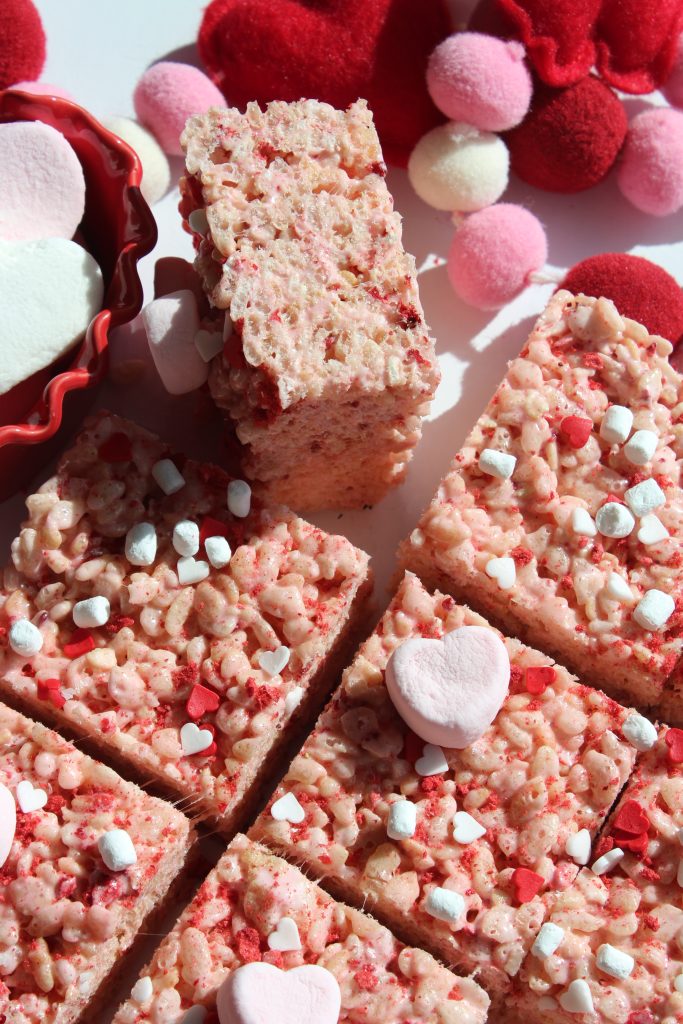 Rice Krispies treats decorated with pink and white marshmallows, red heart sprinkles, and surrounded by fluffy pom-poms and heart-shaped decorations. A bowl of marshmallows is visible in the background. The treats are cut into squares.