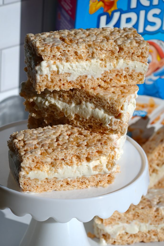 Three large Rice Krispies treats sandwiches with a creamy filling are stacked on a white cake stand. A cereal box is partially visible in the background.
