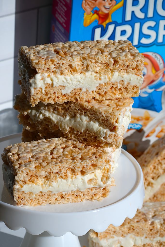 Three ice cream sandwiches made with rice cereal treats are stacked on a white cake stand. A cereal box is partially visible in the background. The sun casts shadows on the treats, highlighting their crispy texture and creamy filling.