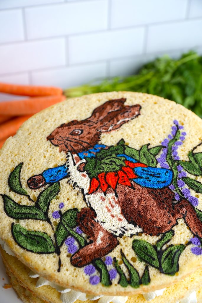 A round cake features an artistic depiction of a rabbit in a blue jacket and red waistcoat, surrounded by green leaves and purple flowers. Carrots are artfully arranged on the white tiled surface, making it one of those fun and easy baking recipes that's also delightful to admire.