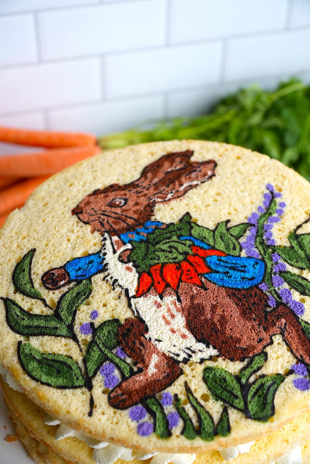 A round cake features an artistic depiction of a rabbit in a blue jacket and red waistcoat, surrounded by green leaves and purple flowers. Carrots are artfully arranged on the white tiled surface, making it one of those fun and easy baking recipes that's also delightful to admire.