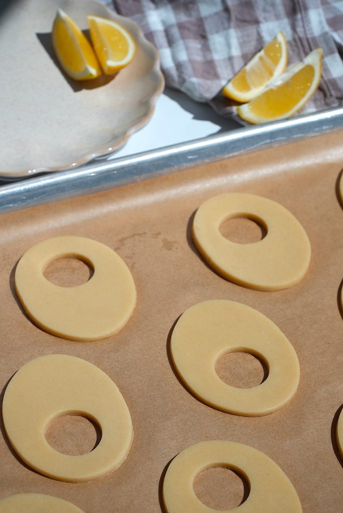 Close-up of unbaked oval-shaped cookie dough with holes in the center on a baking sheet. In the background, a checkered napkin and lemon wedges on a plate are visible.