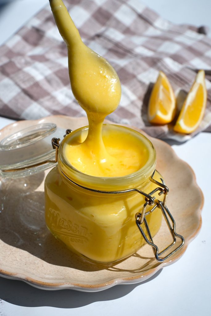 A spoon covered in creamy yellow lemon curd hovers over an open mason jar filled with the curd. The jar sits on a beige plate with lemon wedges and a checkered cloth in the background.