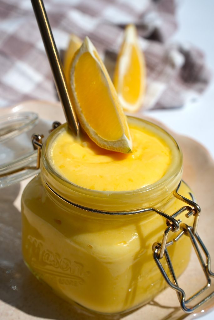 A jar filled with creamy orange mousse is topped with orange slices. A metal spoon is inside the jar. In the background, more orange slices rest on a checkered cloth. The jar sits on a round tray.