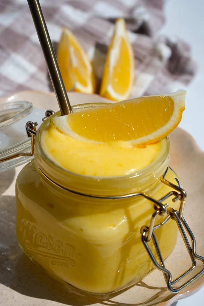 A glass jar filled with creamy lemon mousse, topped with a lemon wedge and a spoon. In the background, three lemon wedges are placed on a checkered cloth. The scene is bathed in natural sunlight.