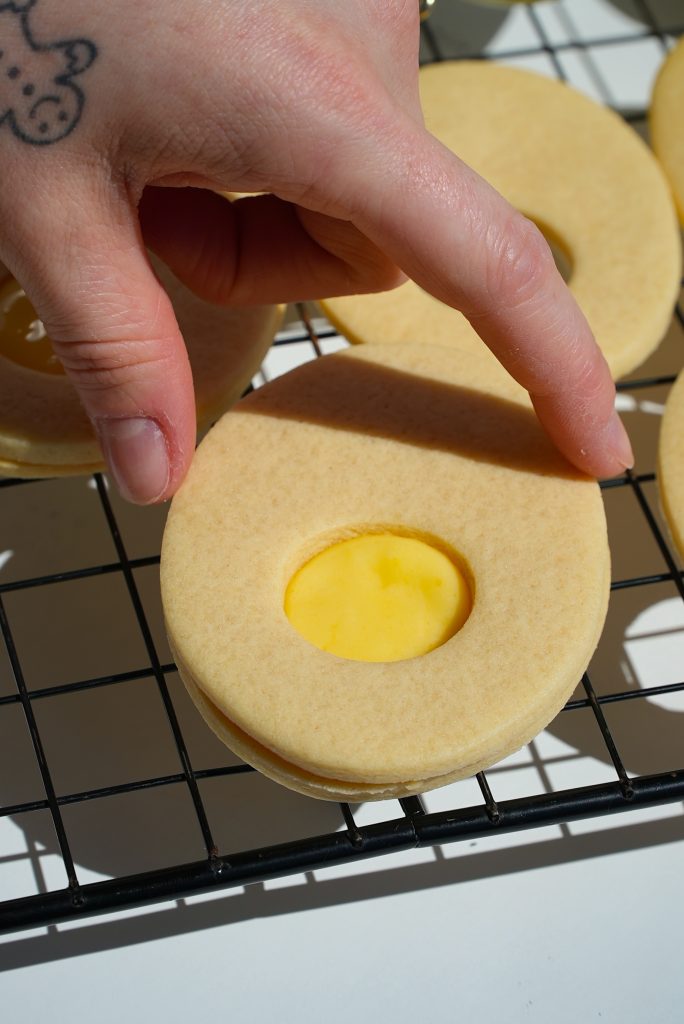 A hand holding a round, unbaked cookie with a smaller circular cutout in the center. It's placed on a black wire rack. Sunlight casts shadows on the surface, and a tattoo is visible on the hand.