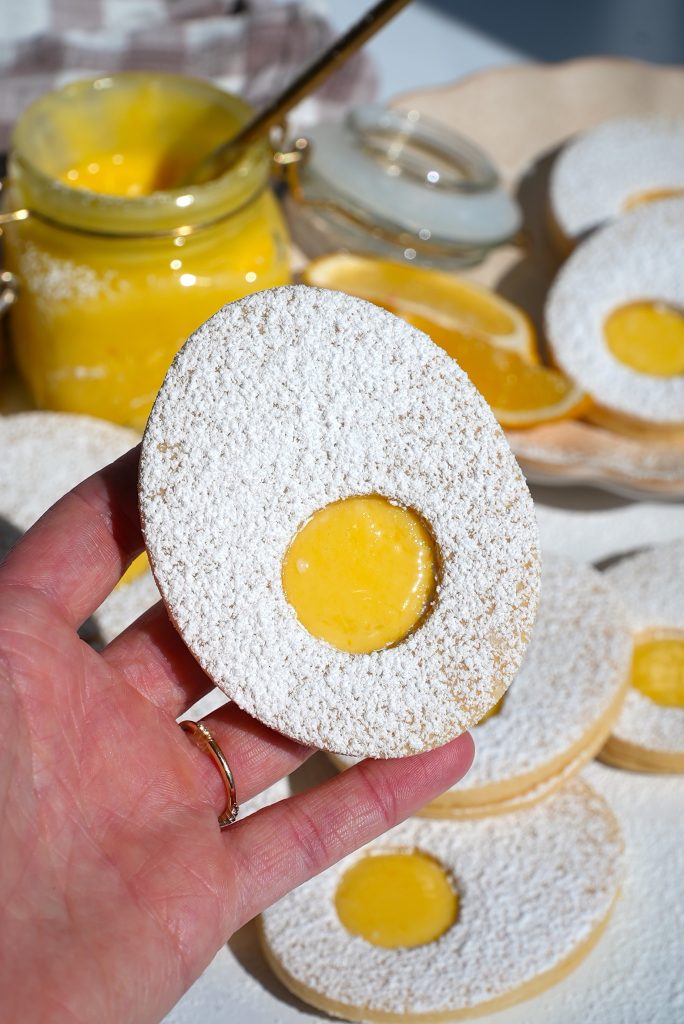 A hand holding a powdered sugar-coated cookie with a yellow filling in the center. A jar of similar yellow filling and more cookies are in the background on a light surface.