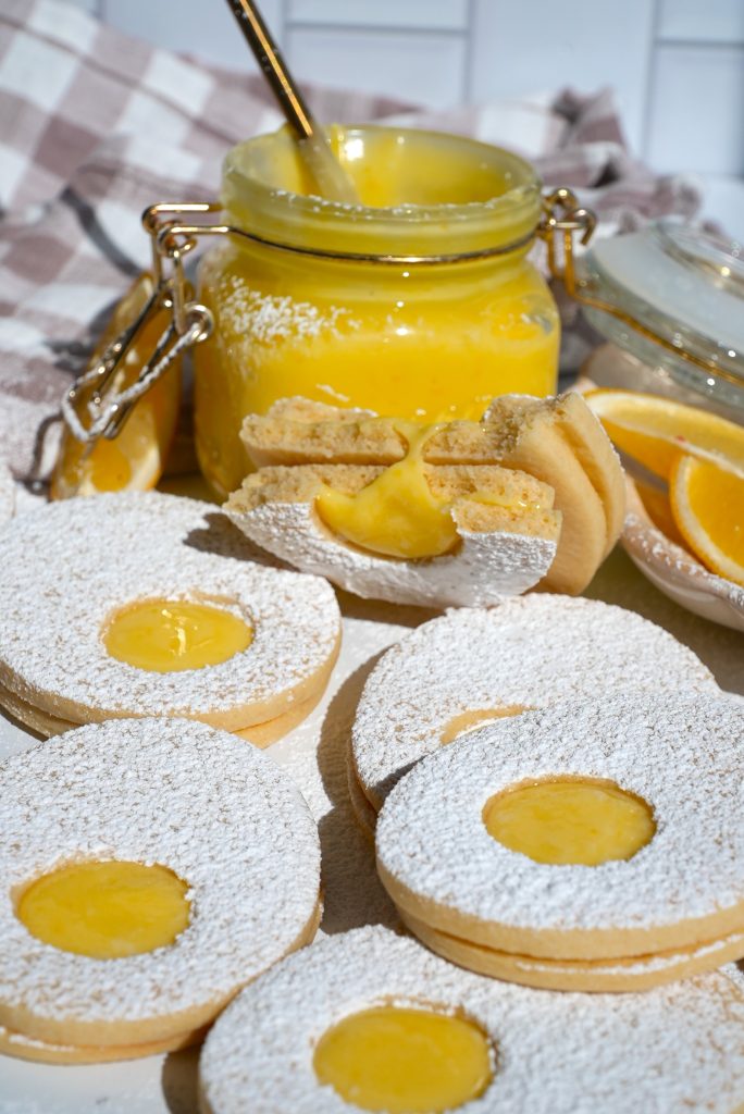Biscuits dusted with powdered sugar and filled with lemon curd are arranged in front of a jar filled with lemon curd. A spoon is inside the jar, and there are lemon slices nearby. A plaid cloth is in the background.