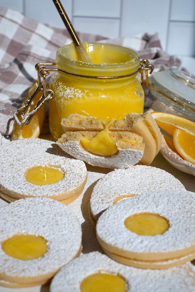 Round cookies with powdered sugar centers filled with yellow lemon curd spread are arranged in the foreground. A jar of lemon curd, topped with a spoon and set on a checkered cloth, is in the background. Slices of lemon are visible nearby.