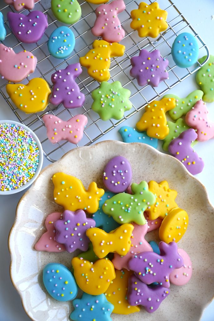 A variety of pastel-colored, animal-shaped cookies with multicolored sprinkles are on a cooling rack and a plate. Nearby, there's a small bowl filled with colorful round sprinkles.