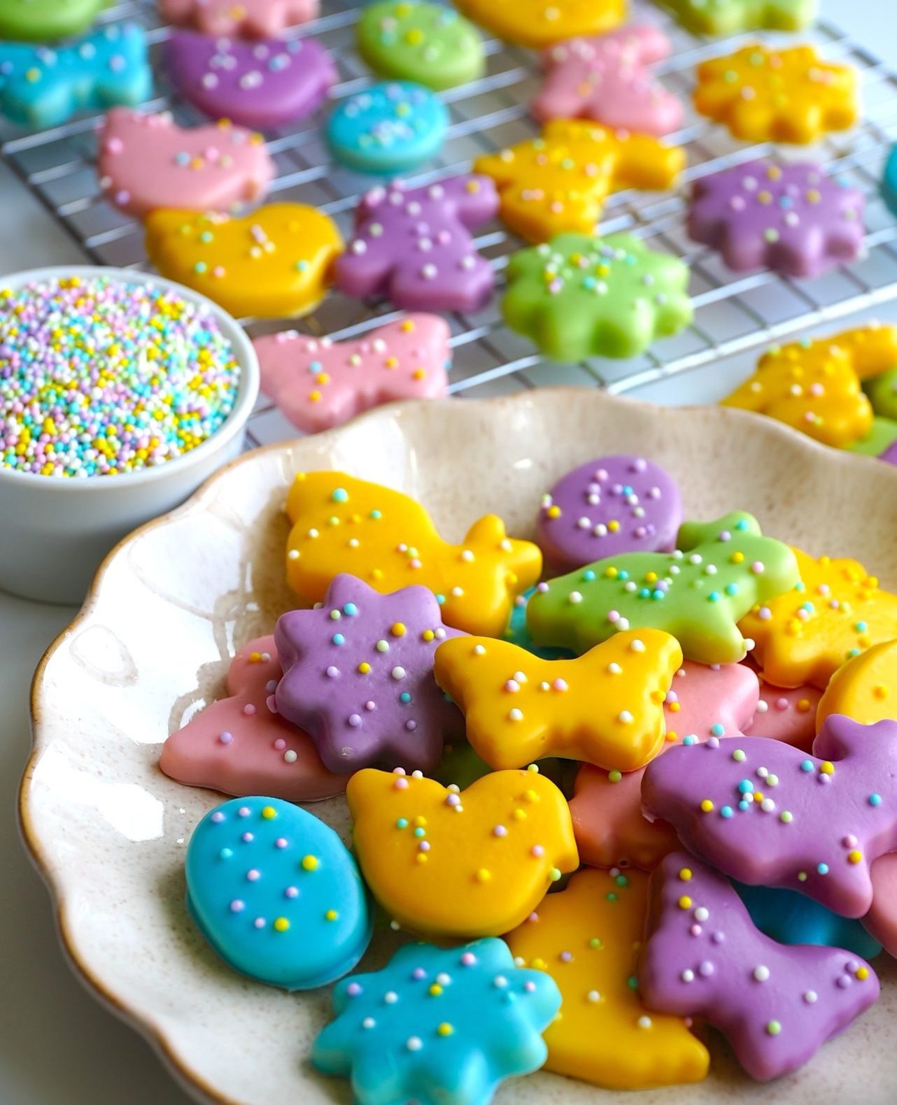 A variety of colorful, animal-shaped frosted cookies with sprinkles are displayed on a wire rack and a white plate. A small bowl of multicolored sprinkles is next to the plate.