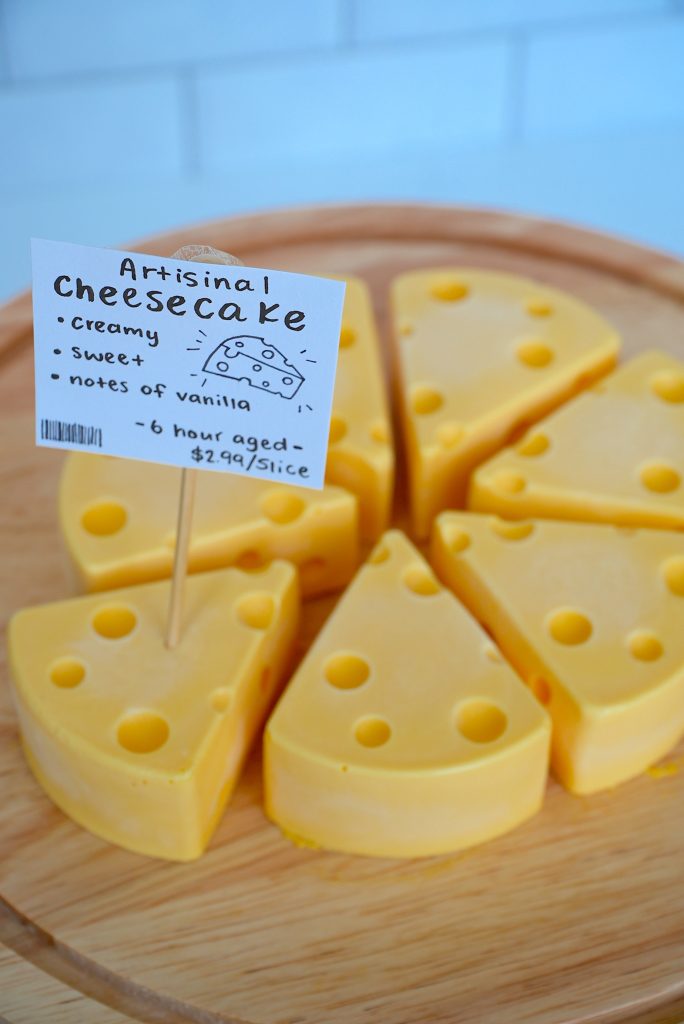 A round cheesecake cut into eight slices, each decorated to look like Swiss cheese. A sign nearby reads “Artisanal Cheesecake: creamy, sweet, notes of vanilla, 6 hour aged, $2.99/slice.”.