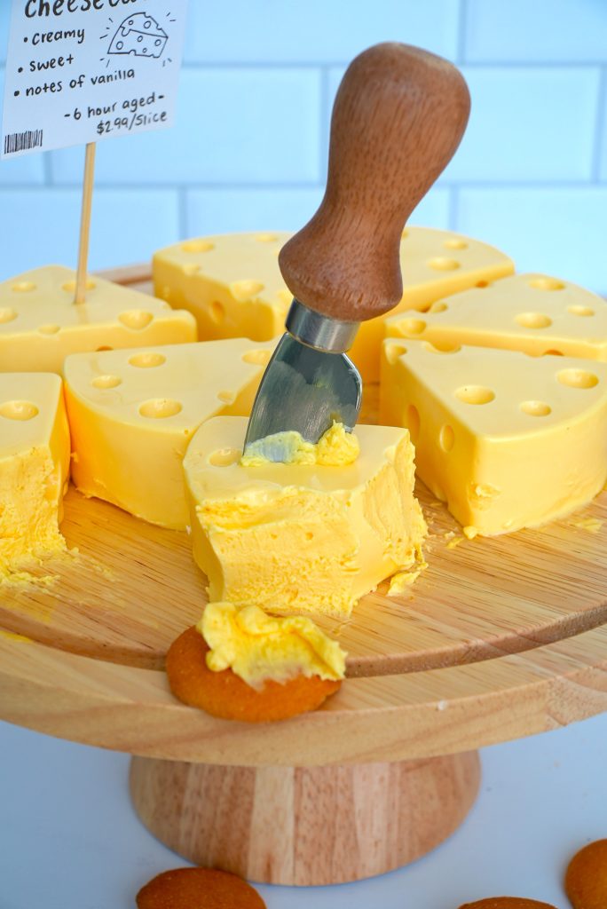 A wooden platter holds slices of creamy yellow cheesecake shaped like Swiss cheese wedges. A cheese knife with a wooden handle is spreading cheesecake onto a cookie. A small sign describes the cheesecake’s flavor and price.