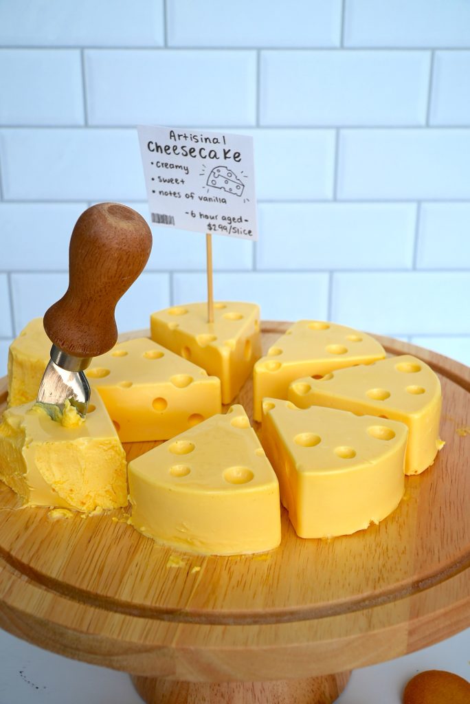 A round wooden board holds wedges of cheesecake shaped like Swiss cheese, arranged in a circle. A small sign describes it as "Artisanal Cheesecake." A serving tool is cutting one slice.