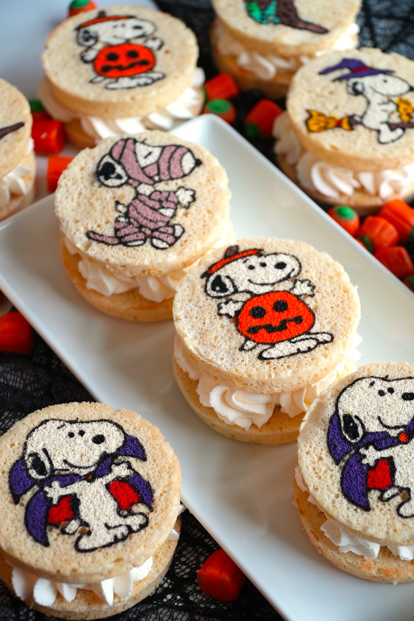Round sandwich cookies with piped white filling, topped with edible images of Snoopy in Halloween costumes—mummy, vampire, witch, and pumpkin—are arranged on a white platter, surrounded by candy corn.